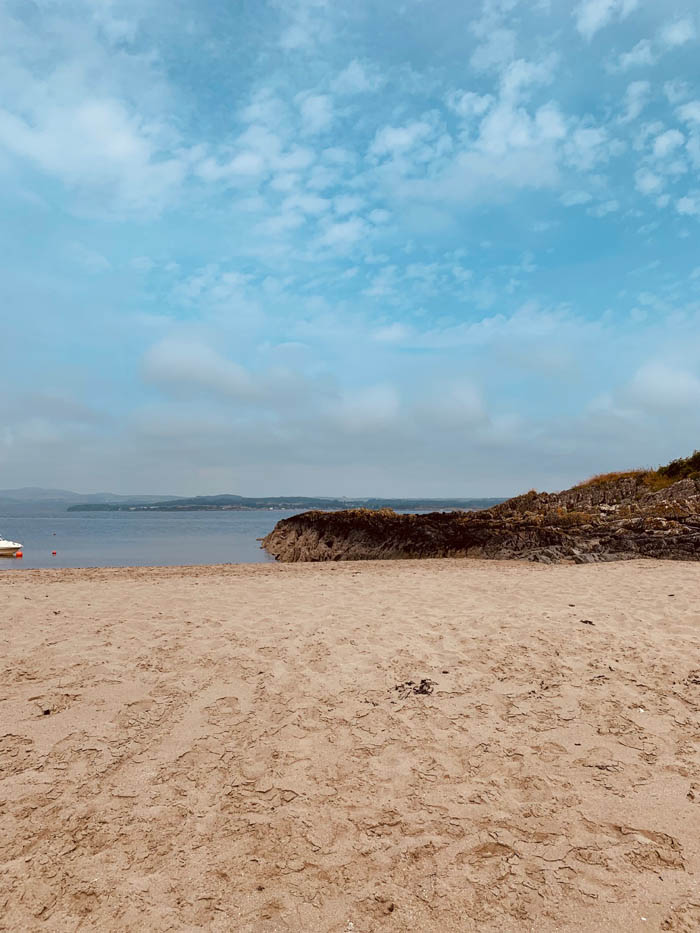 The sand of Mossyard beach. The sand is dimpled, having clearly been well-trod and there is a little white rowing boat partially visible in the left middle of the shot. Beyond the sand are rocky outcrops.